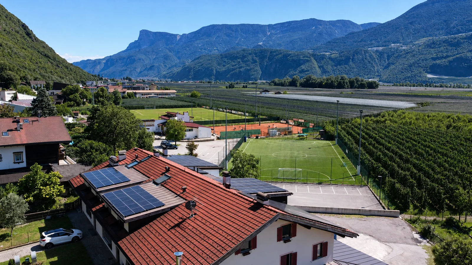 Drohnenaufnahme einer Photovoltaikanlage auf rotem Ziegeldach mit Sportplätzen, Obstgärten und Südtiroler Bergen